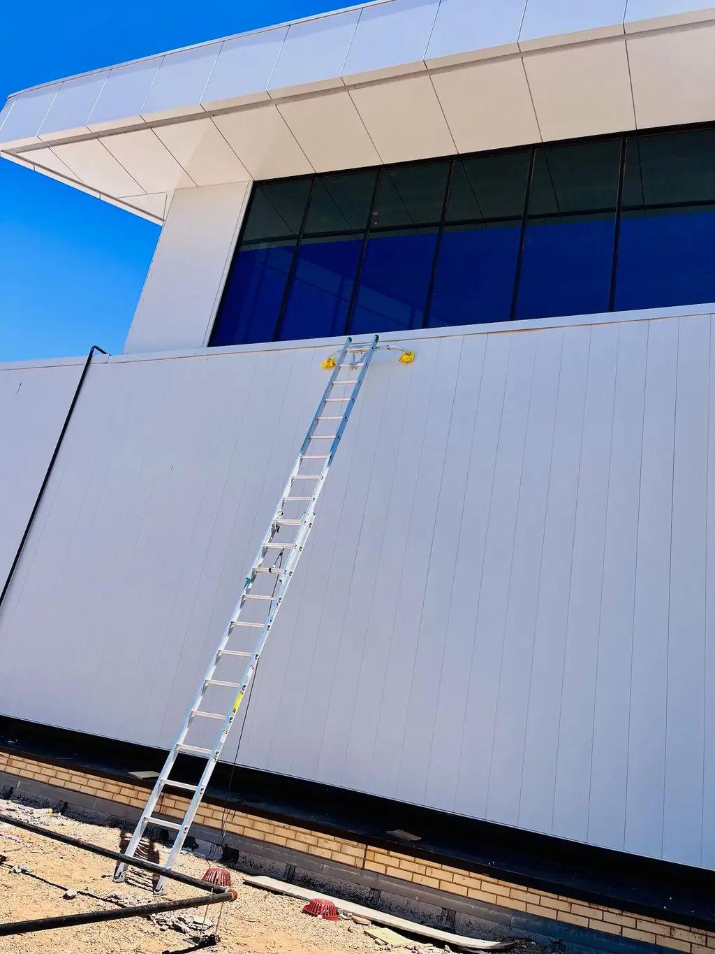 An extension ladder and water-fed pole against a modern metal-panel commercial building with blue glass clerestory windows in Lubbock