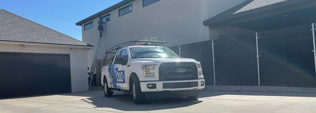 A Caprock Window Cleaning pickup with a ladder rack parked outside a modern white stucco home in Lubbock while a crew member works up a ladder