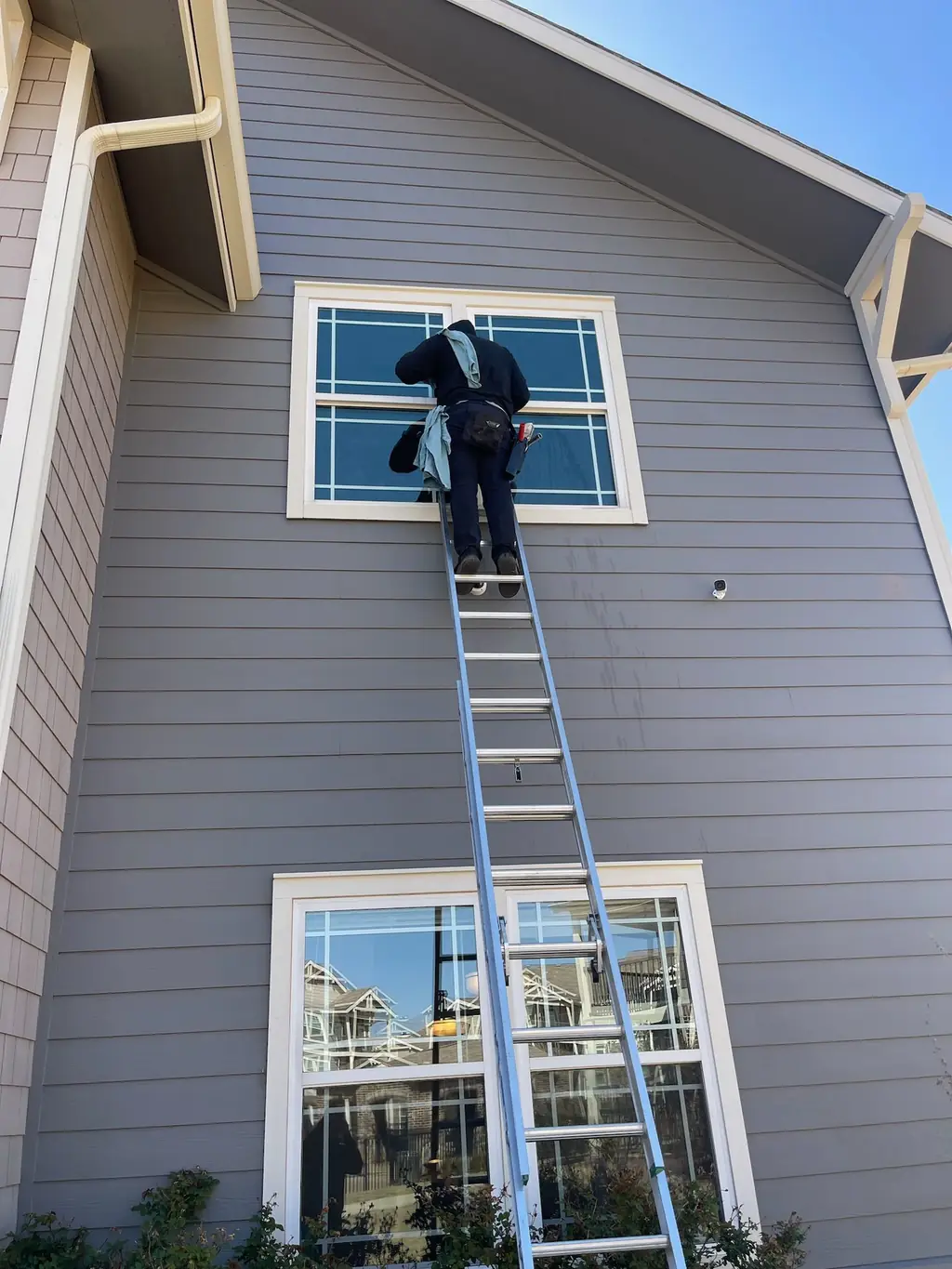 A Caprock crew member up a full extension ladder cleaning a second-storey window on a grey clapboard home under a blue sky