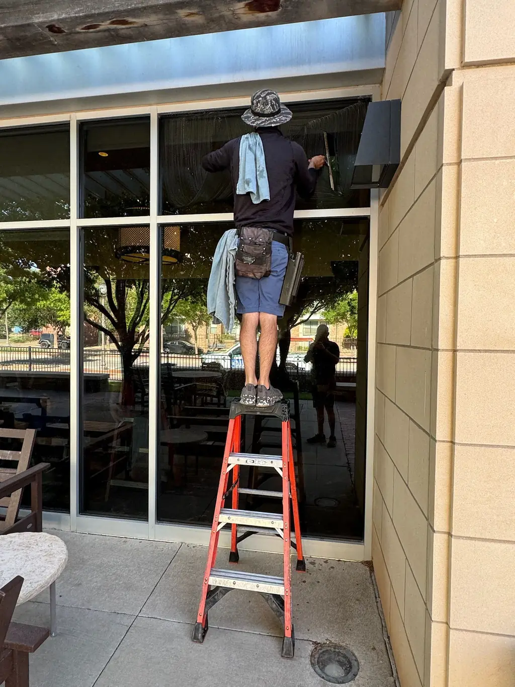 A Caprock crew member on an orange step ladder cleaning tall storefront glass at a commercial patio in Lubbock